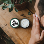 "Open tin of Forest Fresh Whipped Tallow Cream by Lulu's Untainted Tallow, formulated for men with combination skin, available in 60ml and 30ml sizes. The tin sits on a wooden surface with a man applying cream to his face. The label features a vintage-inspired design with a portrait of a woman, a dark green border, and the words 'UNTAINTED TALLOW.' A potted plant and dried foliage add a natural, rugged feel."
