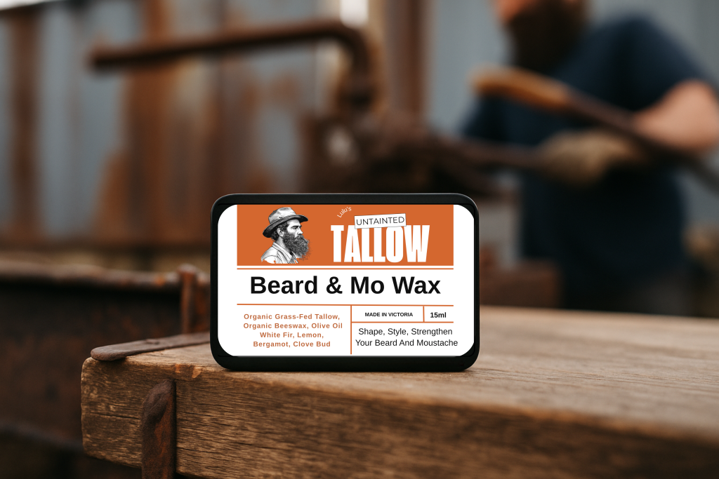 A rectangular black tin of “Lulu’s Untainted Tallow Beard & Mo Wax” sits on a rustic wooden surface in an industrial workshop setting. The label features an orange and white design with a bearded man illustration, listing ingredients such as organic grass-fed tallow, beeswax, olive oil, white fir, lemon, bergamot, and clove bud. A blurred background shows a man working with metal tools, creating a rugged, masculine atmosphere.